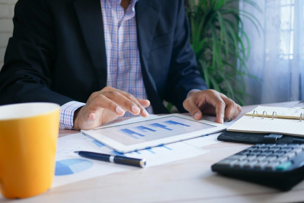 A person in a suit reviews a bar graph on a tablet at a desk with a calculator, notebook, and papers.